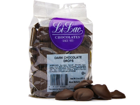A clear cellophane bag of dark chocolate drops sits with a few chocolate drops scattered on the table next to it.