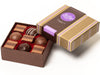 A small, square box containing nine assorted truffles. The box base and lid are gold with narrow purple stripes. Inside are four chocolate ganache truffles in assorted milk and dark chocolate flavors. There are also five hazelnut truffle squares.