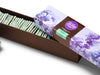 A  rectangular box that is long and narrow. The box has a brown base and a purple lid with watercolor lilac motifs. The box contains a neat row of chocolate mint squares. For each piece a bright green, fresh-mint filling is sandwiched between two delicate layers of dark chocolate. 