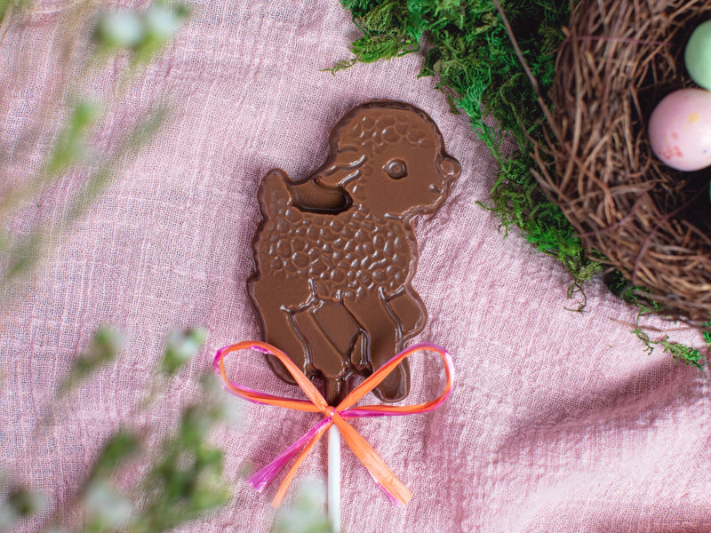 A chocolate lamb pop siting on a pink cloth with a birds next and eggs next to it.