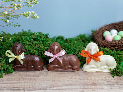 Three chocolate lambs sitting on a board with an Easter scene in the background