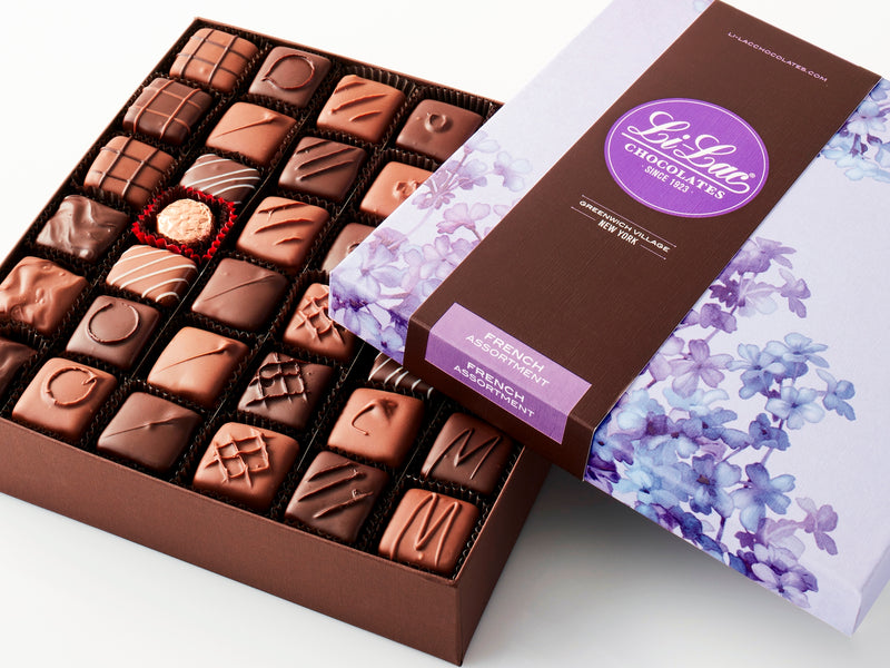 An open box displaying the chocolates inside with the cover partially covering the  top showing a floral lilac pattern on the box top