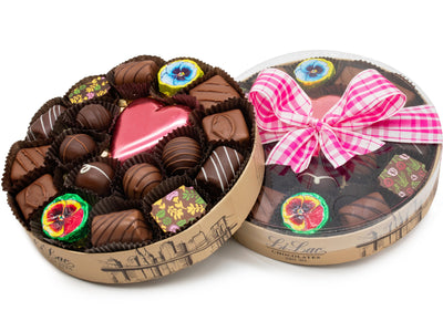 A round box with a clear lid is tied with a springtime ribbon. The box contains 20 pieces of assorted chocolates including a large pink foiled chocolate heart and foiled chocolate pansies.