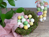 colorful pastel malted eggs artfully arranged in a nest, with a bag of eggs standing behind it.