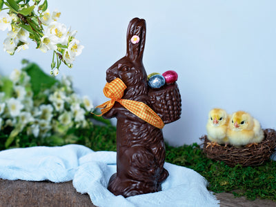 a large chocolate bunny carrying a basket on his back with three foiled eggs.