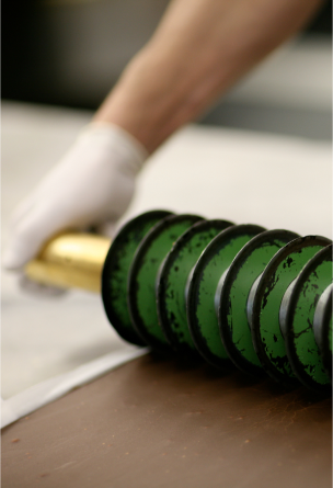 A chocolate maker rolls a long chocolate cutter along a slab of chocolate