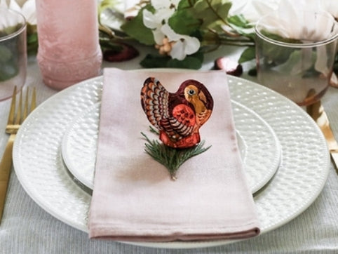 A Dark Chocolate Foiled Turkey sits on a sprig of evergreen decorating a placesetting on a holiday table.