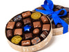 A round box with a clear lid is tied with a blue ribbon. The box contains 20 pieces of assorted chocolates including pieces of gold foiled gelt.