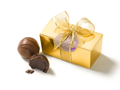 Two assorted gourmet chocolate ganache truffles sit next to a small rectangular gold foil cardstock box. The box is sealed with a circular purple Li-Lac Chocolates sticker and tied with a gold ribbon bow.