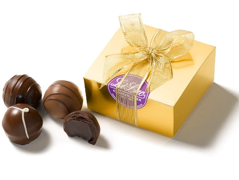 Four assorted gourmet chocolate ganache truffles sit next to a small square gold foil cardstock box. The box is sealed with a circular purple Li-Lac Chocolates sticker and tied with a gold ribbon bow.