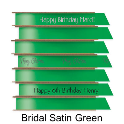 A stack of seven spools of shiny green satin ribbon with custom messages printed on them in different colors. 