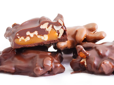 Pecan chews enrobed in milk or dark chocolate are stacked together. One is cut in half revealing the interior of a big dollop of caramel studded with pecans.
