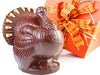 A large chocolate molded turkey stands next to a decorative square box tied with a ribbon.  The turkey has candy corn decorations on its fanned out tail.