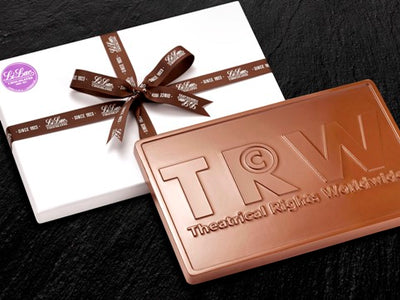 A large 1 pound chocolate bar has a business logo T R W embossed onto it.  It leans on a white gift box tied with a brown Li-Lac Chocolates branded ribbon.