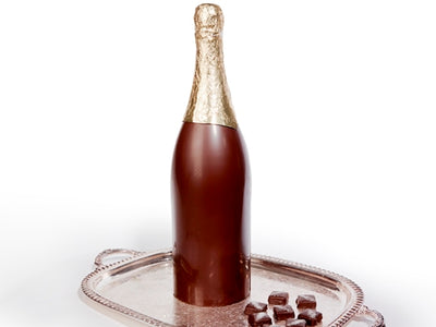 A large, magnumm sized dark chocolate champagne bottle stands on a silver platter. The bottle has a gold foiled top to look like a real bottle of champagne.