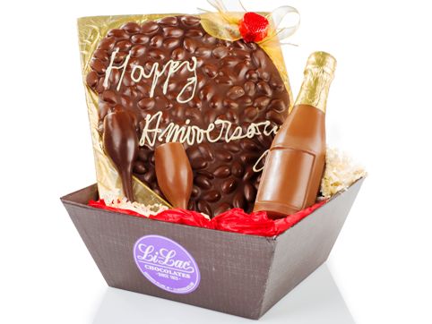 A low, dark brown, square box holds one milk chocolate and one dark chocolate champagne glass, a 7” Chocolate champagne bottle with a gold foil top. A large oval sheet of chocolate almond bark sits upright on a gold cardstock base. The greeting "Happy Anniversary" is hand written on the sheet of almond bark in white chocolate.