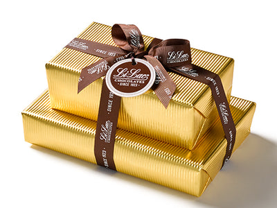 Two boxes of chocolate have been beautifully gift wrapped in gold foil paper. They are stacked on top of each other and tied with a dark brown Li-Lac Chocolates logo printed ribbon with a round gift tag.