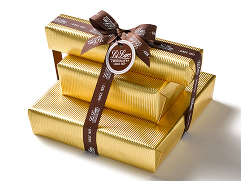 Three boxes of chocolate have been beautifully gift wrapped in gold foil paper. They are stacked on top of each other and tied together with a dark brown Li-Lac Chocolates logo printed ribbon with a round gift tag.
