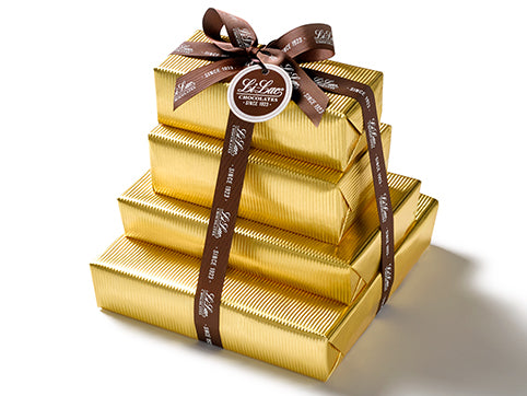Four boxes of chocolate have been beautifully gift wrapped in gold foil paper. They are stacked on top of each other and tied together with a dark brown Li-Lac Chocolates logo printed ribbon with a round gift tag.