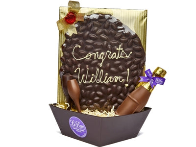 The words "Congrats William" are piped onto an oval of almond bark in white chocolate sits ina square basket with a chocolate champagne bottle and chocolate champagne flute.