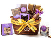 A low, oval wicker basket is tied with a yellow congratulations ribbon. The basket holds one home assortment chocolate box, butter crunch in a purple window box, one boxed chocolate bar, a bag of chocolate espresso beans, a thank you bar in a gold box with a brown ribbon, salted caramels in a purple window box, a 7” chocolate champagne bottle.