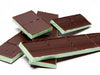 Strips of French Mint Bars are stacked together. Each piece has a bright green, fresh-mint filling that is sandwiched between two delicate layers of dark chocolate. Each strip is scored, intended to break into four squares.