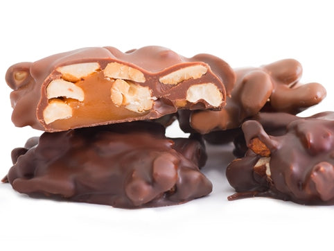 Cashew chews enrobed in milk or dark chocolate are stacked together. One is cut in half revealing the interior of a big dollop of caramel studded with cashews.