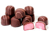 Mounds of raspberry soft cream are enrobed in dark chocolate with pink drizzle on top. One of the raspberry creams is cut in half reveling a soft pink raspberry cream center. 