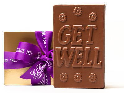 A large rectangular chocolate bar has the words 'Get Well' written on it. It comes in a gold gift box with a purple ribbon.