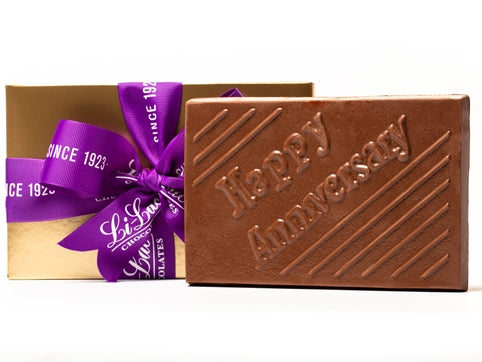 A large rectangular chocolate abr has the words "Happy Anniversary" molded onto the front. It comes in a gold gift box tied with a purple ribbon.