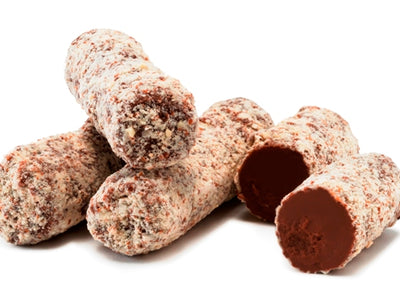 Little cylindrical rolls of chocolate hazelnut ganache are lined up together. They are covered in milk chocolate and coated with crushed almonds. One log is cut in half revealing the soft ganache interior.