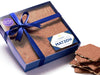 Sheets of matzoh cracker are enrobed in chocolate and sit inside a blue gift box with a clear lid and tied with a blue ribbon.