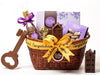 A low, oval wicker basket is tied with a yellow congratulations ribbon. The basket holds one life-sized chocolate champagne bottle with a gold foil top, a Home Assortment box of chocolates, one tall townhouse, one small townhouse, two chocolate keys and a purple window box with butter crunch.