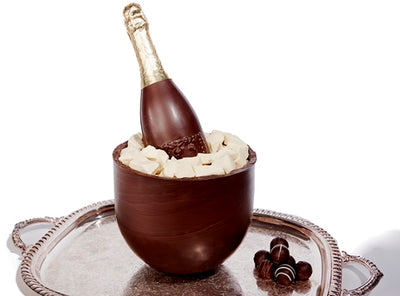 A chocolate champagne bottle, sits inside a chocolate bucket. The bucket is filled with white chocolate ice cubes. 