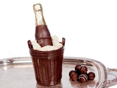 A chocolate champagne bottle, sits inside a chocolate bucket. The bucket is filled with white chocolate ice cubes. 