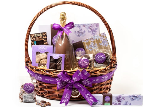 A low, oval wicker basket with a rounded handle is tied with a purple Li-Lac Chocolates ribbon. The basket holds ten beautiful chocolate gifts.