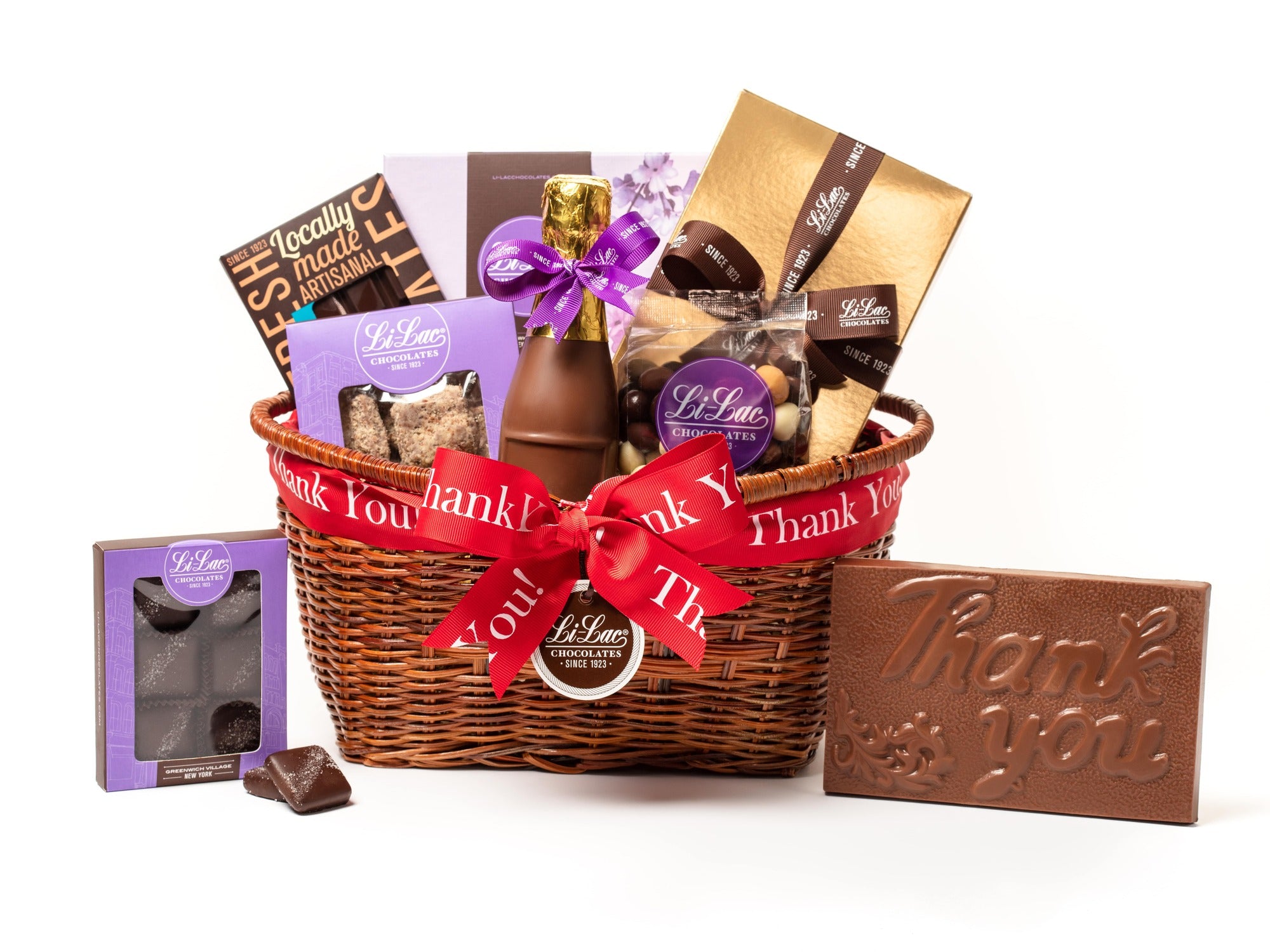 A low, oval wicker basket is tied with a red thank you ribbon. The basket holds one home assortment chocolate box, butter crunch in a purple window box, one boxed chocolate bar, a bag of chocolate espresso beans, a thank you bar in a gold box with a brown ribbon, salted caramels in a purple window box, a 7” chocolate champagne bottle.