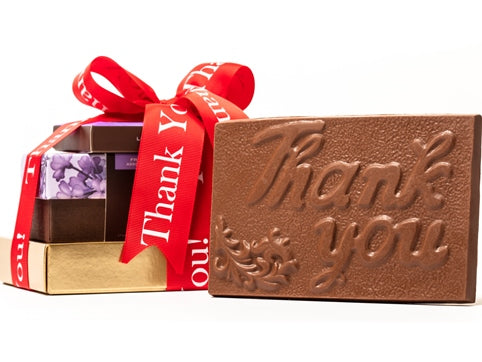 A Thank You chocolate bar in a gold box is stacked with a French Assortment gift box and a window box of caramels all tied together with a red "Thank You" printed ribbon.