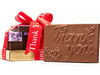 A Thank You chocolate bar in a gold box is stacked with a French Assortment gift box and a window box of caramels all tied together with a red "Thank You" printed ribbon.