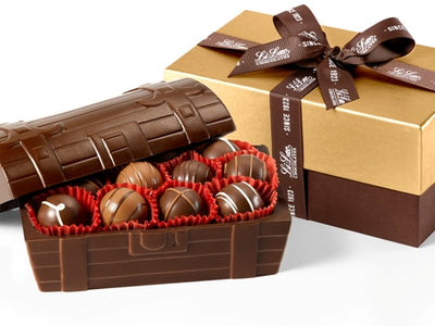 A treasure chest made of molded chocolate sits with the removeable lid half off. Inside the chest are nestled an assortment of 20 gourmet truffles. A rectangular gift box sits behind the treasure chest. The base of the box is dark brown and the lid of the box is gold. It is tied with a dark brown Li-Lac Chocolates branded ribbon.