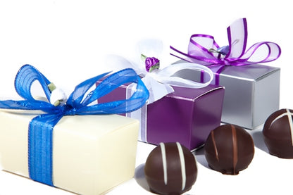 Three assorted gourmet chocolate ganache truffles sit next to three small rectangular cardstock boxes in different colors. The boxes are tied with different colored ribbon bow. 