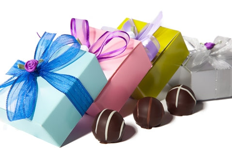 Three assorted gourmet chocolate ganache truffles sit next to three small square cardstock boxes in different colors. The boxes are tied with different colored ribbon bows.