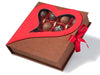 A card box that opens like a book is tied closed with a red ribbon. There is a heart shaped window on the cover where you can see the truffles inside.