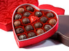 Nineteen assorted chocolates sit inside a heart shaped box. The square, outer box stands nearby.